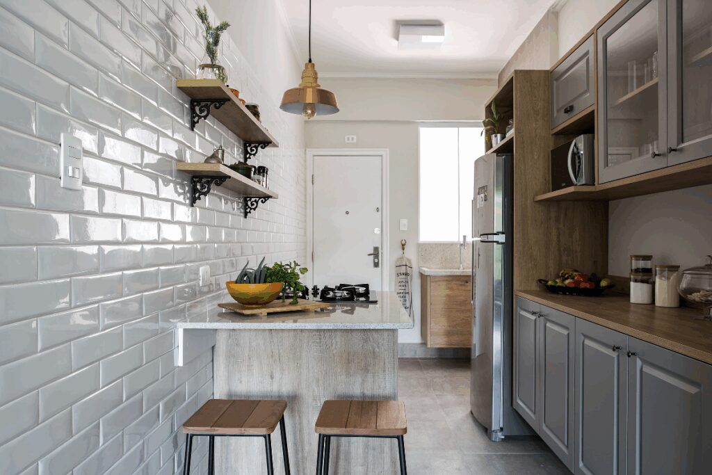 Cozinha com armários em madeira e portas na cor cinza, com bancada de mármore com cooktop e espaço para comer com dois bancos de ferro com assento em madeira e prateleiras na parede de piso branco