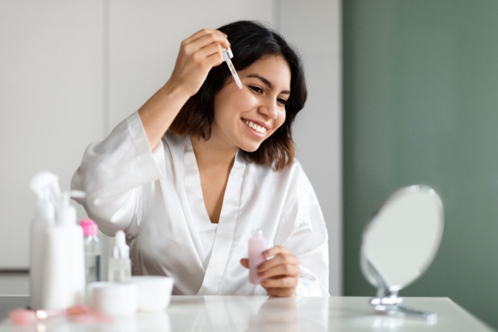Mulher sorrindo, se olhando em um espelho e passando sÃ©rum no rosto