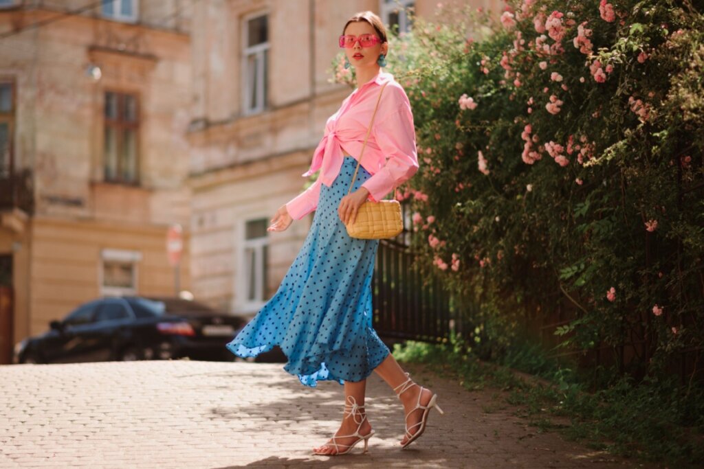 Mulher com o cabelo preso usando camisa e óculos de sol rosa com saia azul de bolinhas pretas e sandália de tiras brancas em jardim