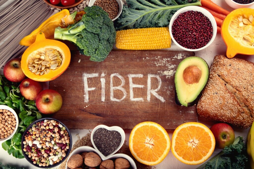 Mesa de madeira, onde estÃ¡ escrito fiber com giz branco e diversos alimentos ricos em fibras como laranja, abacate, milho, maÃ§Ã£ e outros