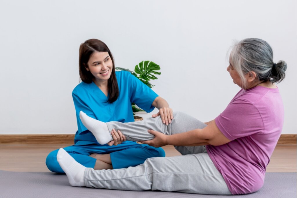 MÃ©dica com o cabelo liso solto, usando um uniforme azul auxiliando a fisioterapia no joelho de uma paciente idosa usando uma camiseta roxa, uma calÃ§a cinza e meia branca