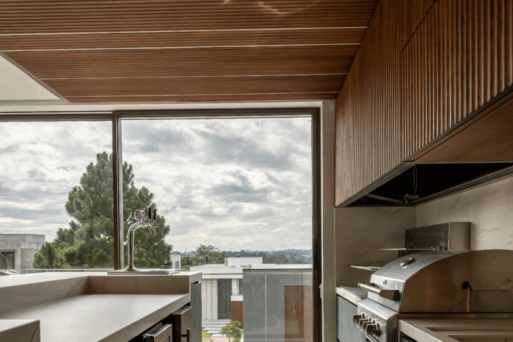 Ãrea gourmet com churrasqueira e forro em madeira e Ã¡rmarios em madeira e bancada de mÃ¡rmore com parede de vidro ao fundo