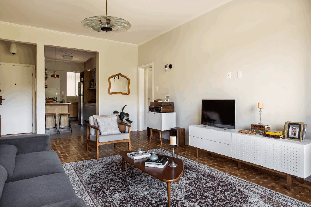 Sala de estar com móvel em branco com TV e cadeira de madeira com estofado branco e sofá preto e tapete estampado no chão