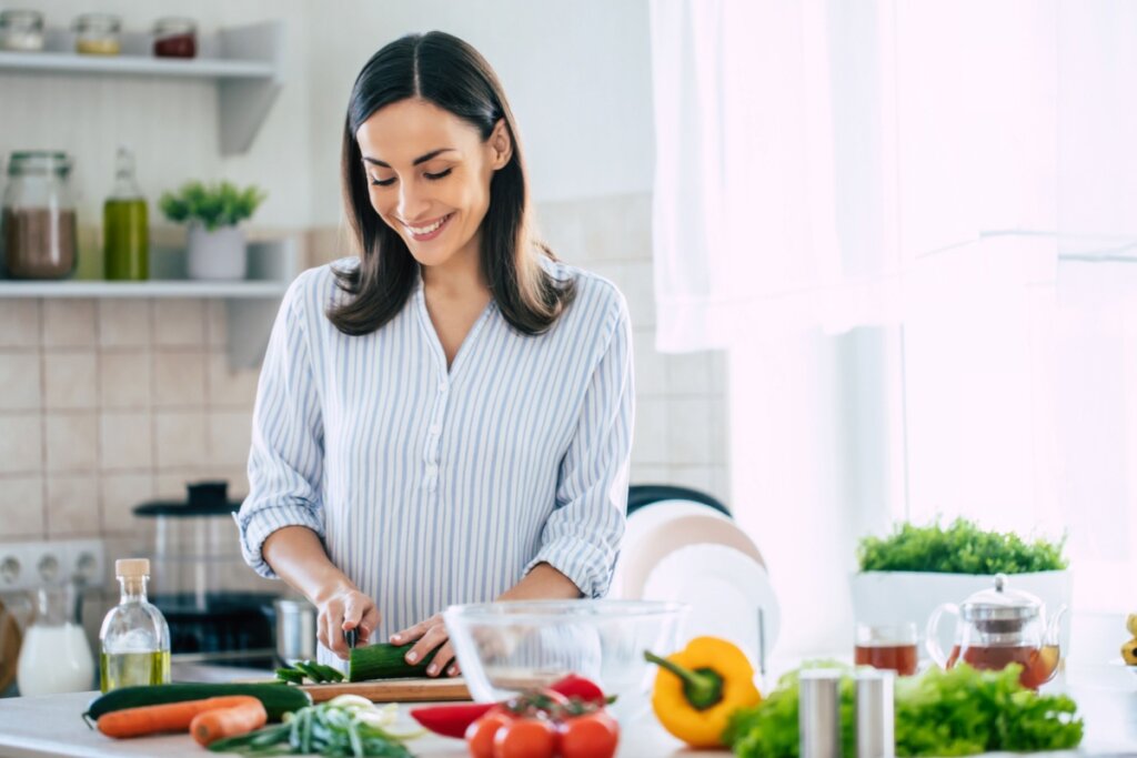 mulher de camisa branca e azul, sorrindo e picando legumes em tÃ¡bua. Ã  frente dela, hÃ¡ diversos alimentos vegetais
