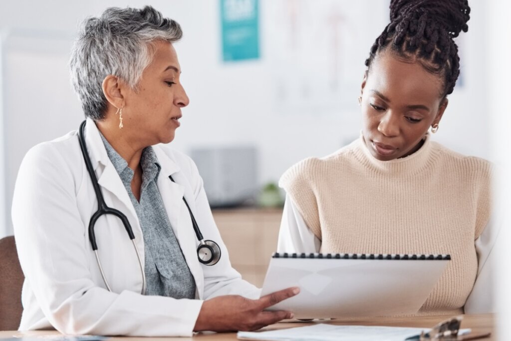 Jovem no consultÃ³rio com mÃ©dica analisando exames. A mÃ©dica tem o cabelo curto e grisalho e estÃ¡ usando jaleco branco e camisa cinza e com estetoscÃ³pio no pescoÃ§o e a paciente tem o cabelo tranÃ§ado preso em coque e estÃ¡ usando camisa de manga longa branca e colete bege.