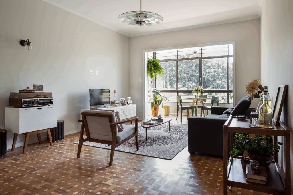 Sala com piso de taco, sofá preto e cadeiras em madeira com estofado branco e móvel com TV branco e paredes pintadas em tons neutros e com janela ampla ao fundo