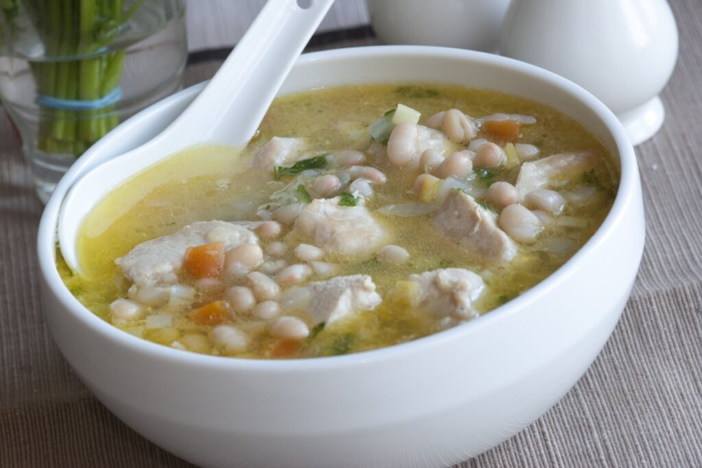 Sopa de feijão-branco com frango servido em tigela de cerâmica branca em cima de mesa de madeira