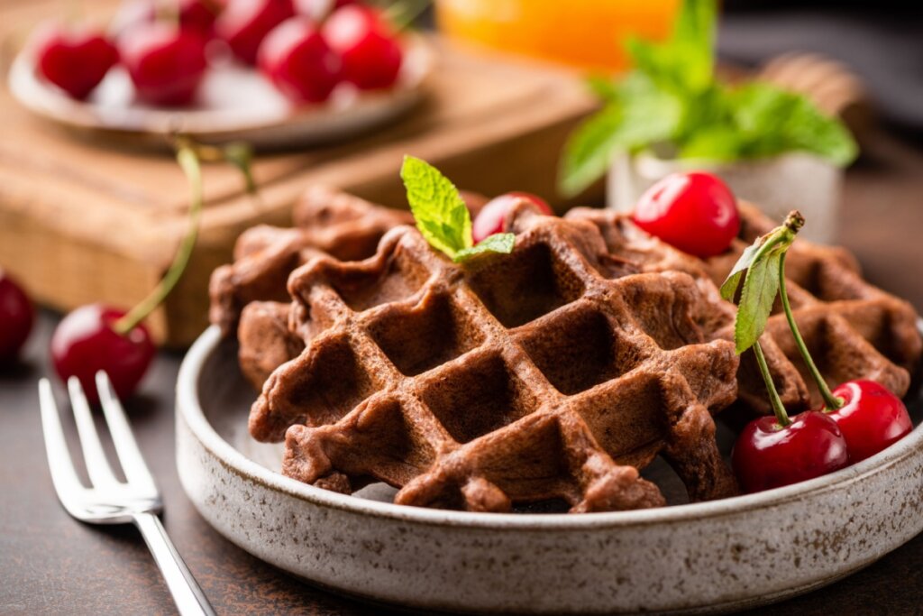 Waffle de chocolate em prato raso de cerÃ¢mica em cima de mesa de madeira, com garfo ao lado e cerejas