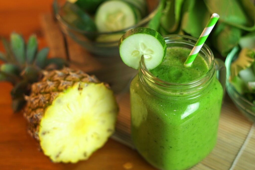 Caneca de vidro com suco de pepino com abacaxi e espinafre em cima de uma mesa com pedaÃ§os de abacaxi, pepino e folhas de espinafre