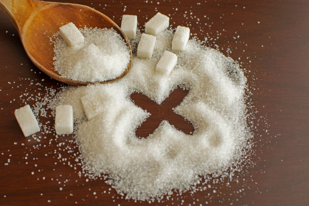 AÃ§Ãºcar esparramado em mesa de madeira com o desenho de um X e com diversos cubos de aÃ§Ãºcar ao redor e uma colher de madeira com aÃ§Ãºcar ao lado