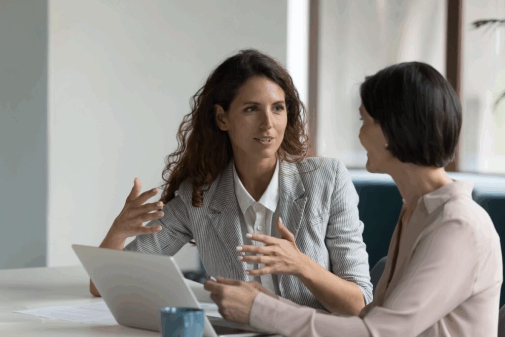Mulher com o cabelo cacheado solto usando camisa de botÃ£o branca e blazer cinza conversando com outra mulher com o cabelo curto e usando camiseta de botÃµes bege