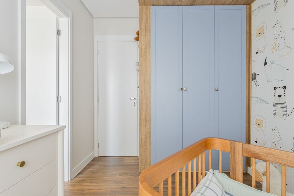 Quarto infantil com parede decorada com papel de parede de girafa, armÃ¡rio de portas azuis, poltrona branca e berÃ§o de madeira