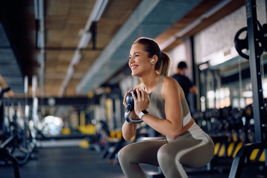 Mulher com o cabelo preso em rabo de cavalo usando roupas de academia cinza com detalhes em branco levantando peso na academia 