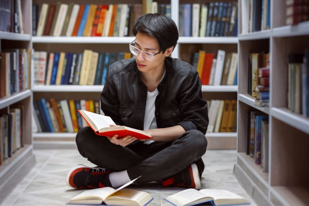 Jovem sentado no chÃ£o de uma biblioteca lendo livro