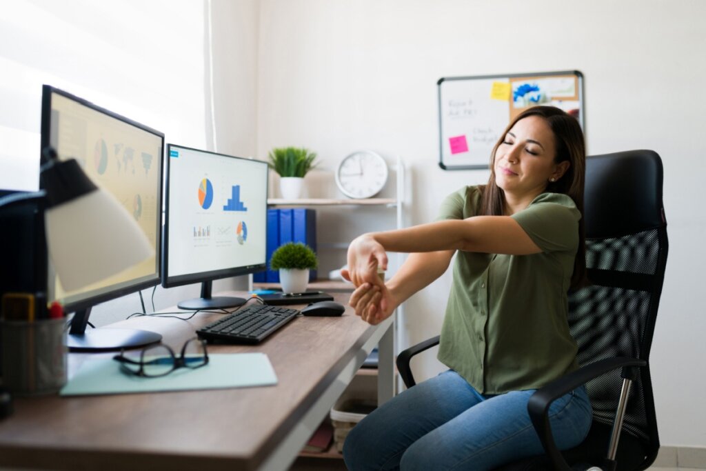 mulher de camiseta verde sentada em frente à duas telas de computador e alongando o braço