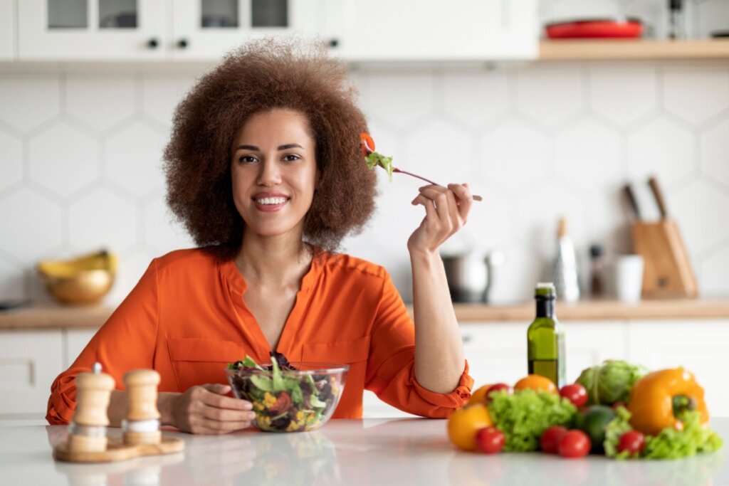 Mulher com o cabelo cacheado curto, usando camisa laranja, sentada à mesa, comendo salada em uma tigela transparente