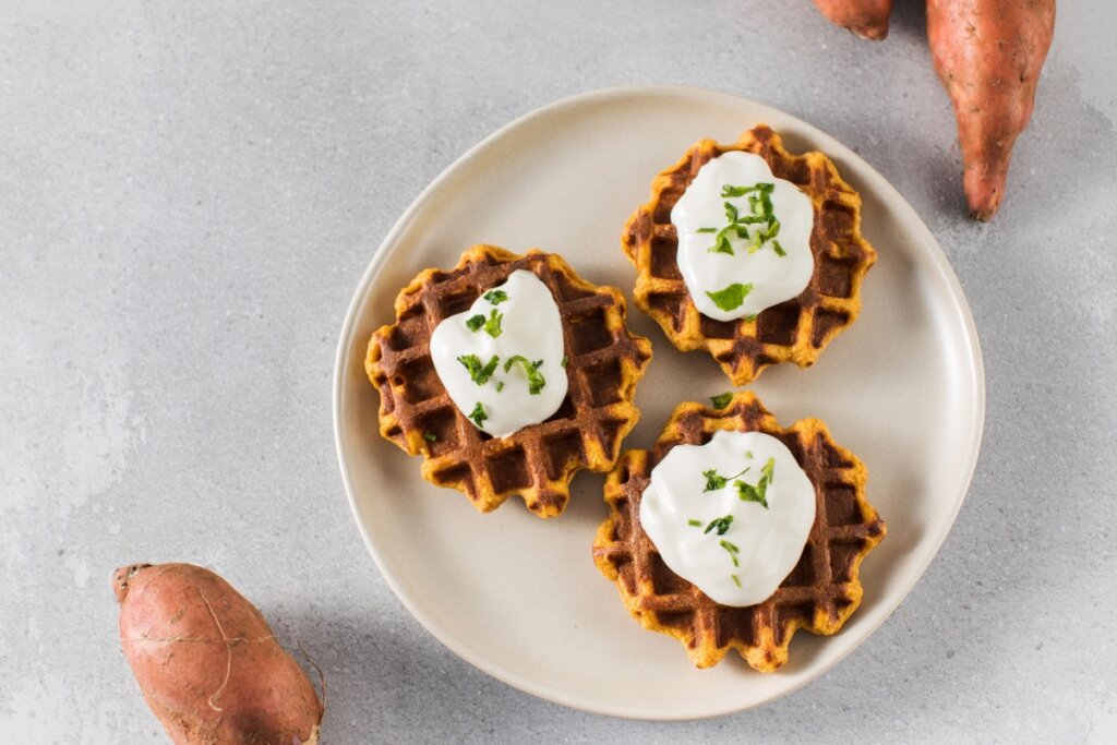 Waffles com creme branco em cima e raspas de limão em prato claro ao lado de batatas-doces