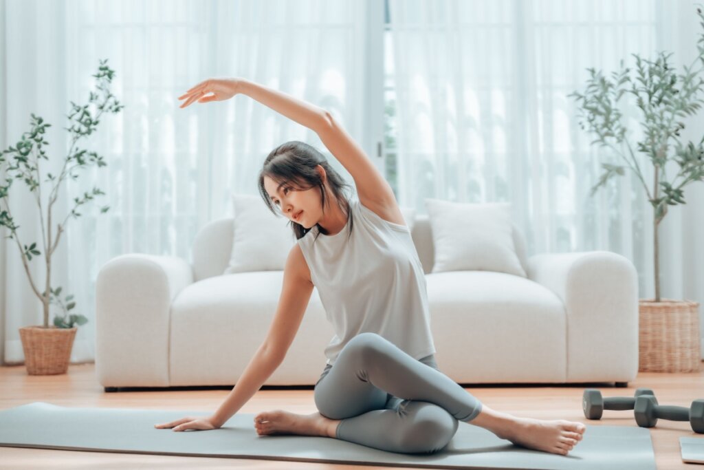 Mulher sentada em tapete realizando alongamento lateral em casa com halteres ao lado