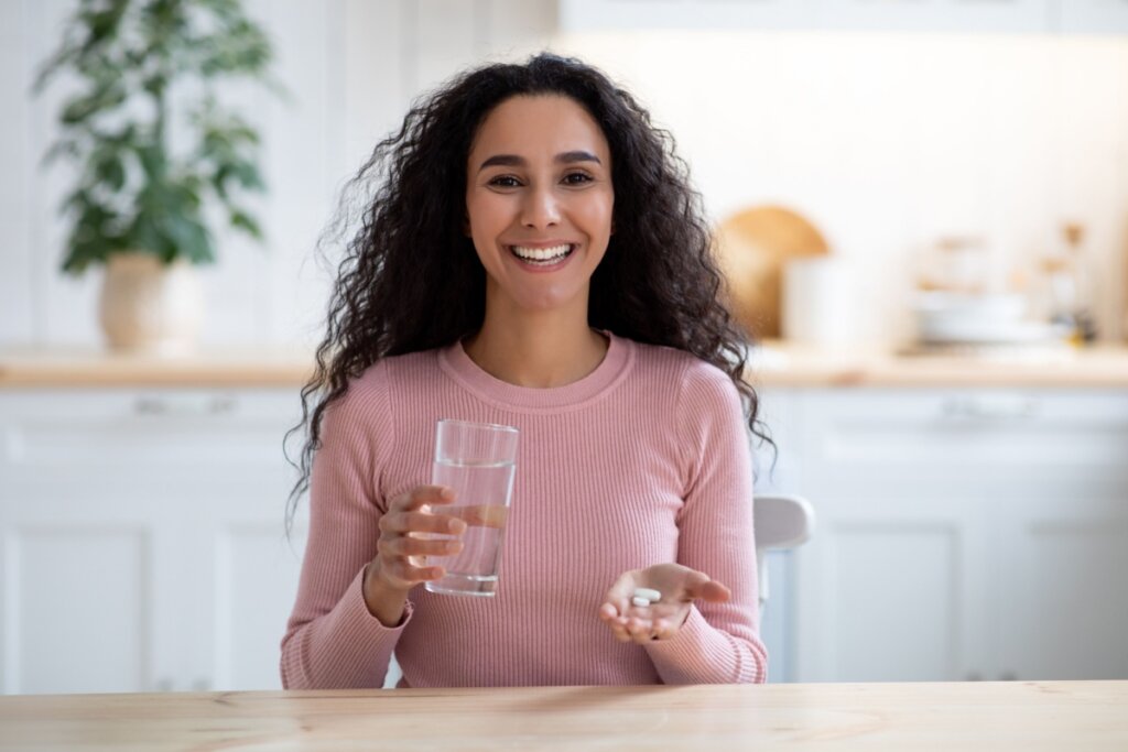 mulher de cabelo cacheado e blusa rosa sorrindo e segurando copo de &aacute;gua e comprimidos 