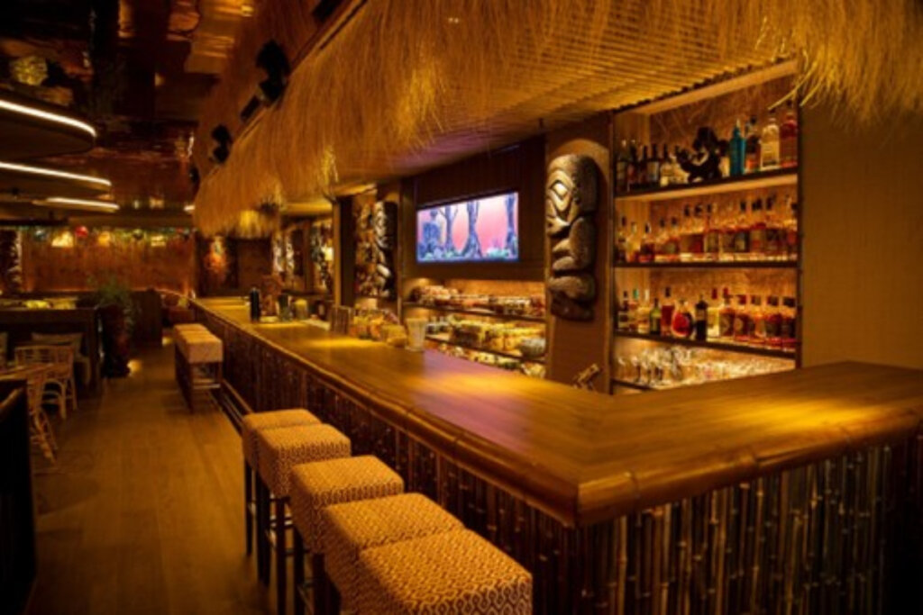 imagem de bar com luz amarela fraca com balcÃ£o grande de madeira e parede com prateleiras com bebidas. bancos em frente ao balcÃ£o