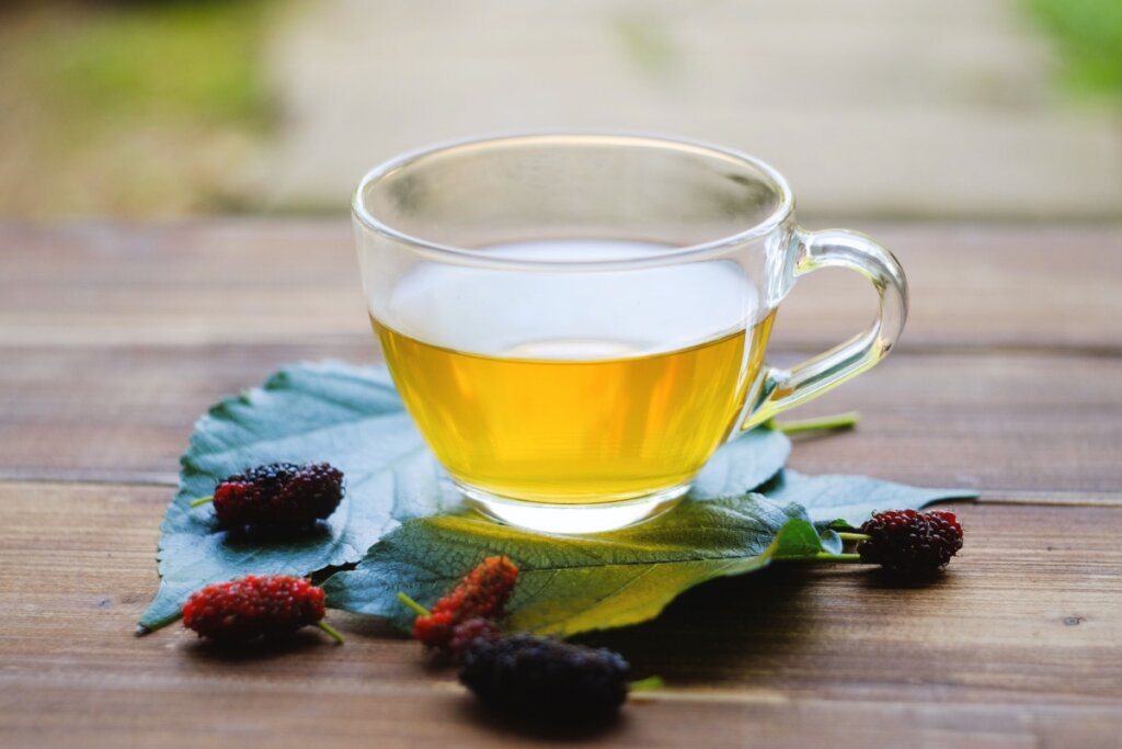 XÃ­cara de vidro transparente com chÃ¡ de amora sobre folhas verdes e frutas de amora fresca