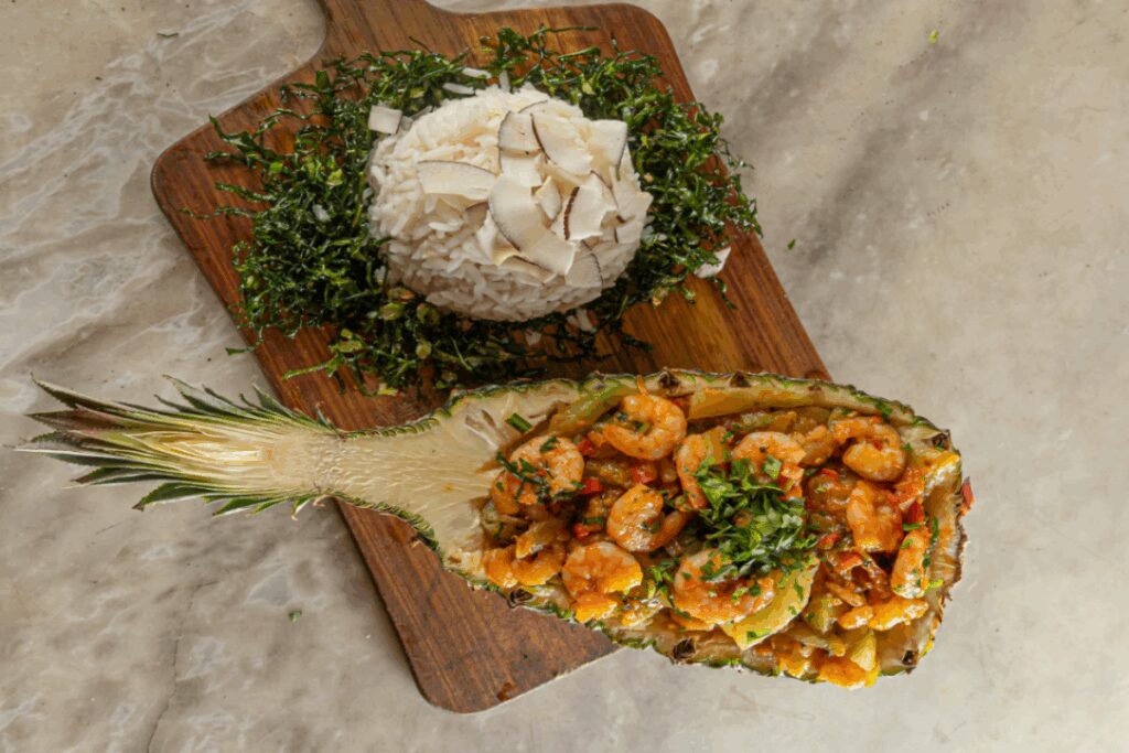 Ensopado de camarão no abacaxi com arroz de coco e couve crispy servido em tábua de madeira em cima de mesa de pedra