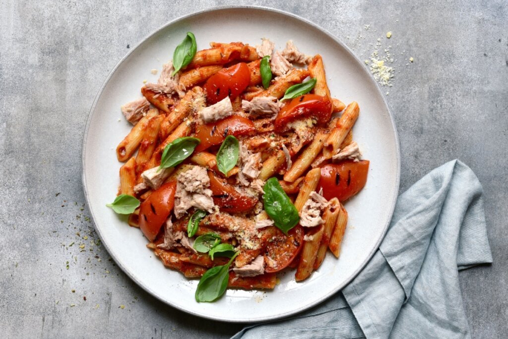 Prato de penne com molho de tomate, pedaços de atum e folhas de manjericão fresco em superfÃcie cinza com pano de cor semelhante ao lado