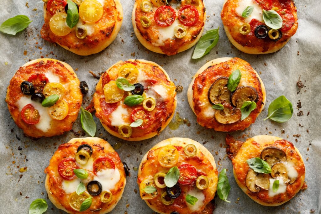 Minipizzas coloridas cobertas com queijo derretido, tomates, azeitonas e rodelas de berinjela, decoradas com folhas frescas de manjericão