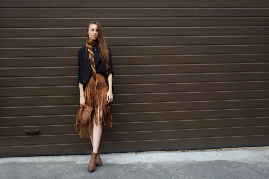 mulher de cabelo longo encostada em parede marrom. ela veste camisa preta, saia marrom com franjas, segura bolsa marrom com franja e usa bota marrom