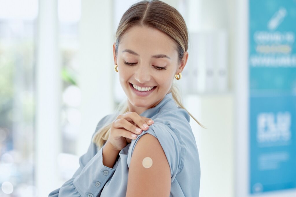 Mulher sorrindo, levantando manga de blusa azul e olhando para curativo de vacina no braÃ§o
