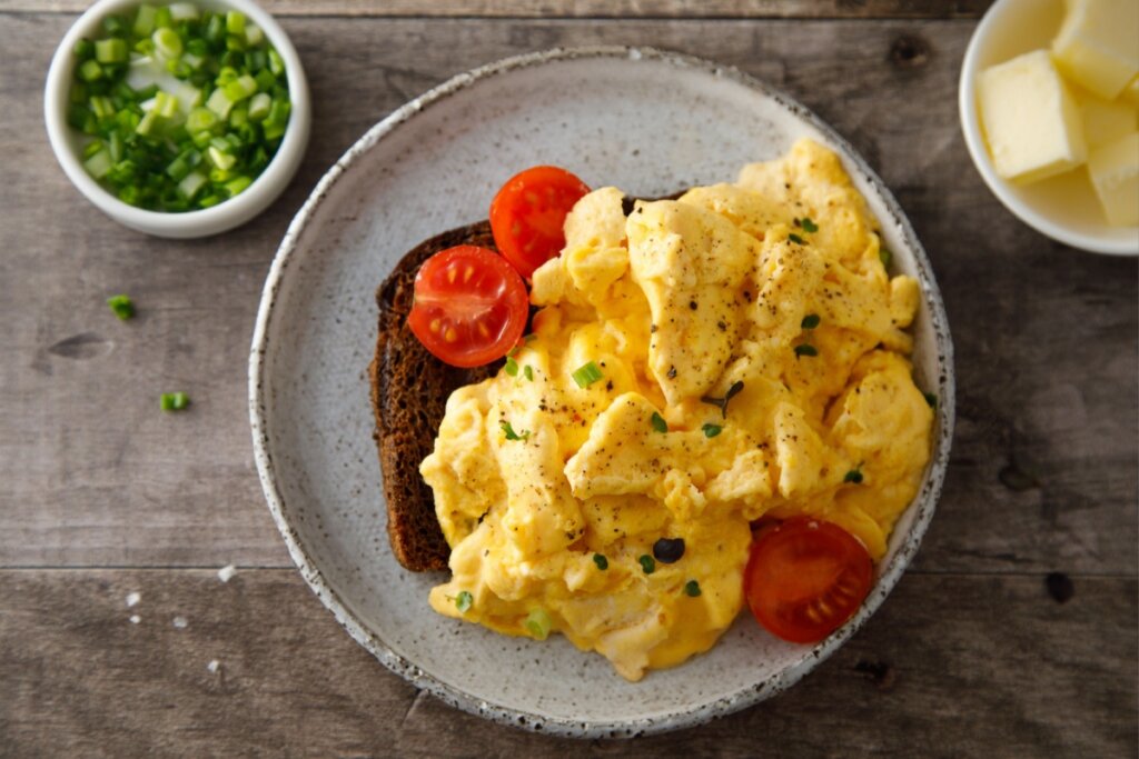 prato cinza com fatia de pão marrom com ovo cremoso mexido e tomate-cereja fatiado