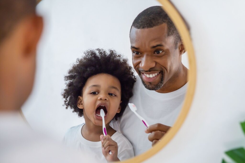 Pai com o cabelo curto e barba, orientando filho com o cabelo cacheado e solto a escovar os dentes em frente ao espelho, ambos estÃ£o usando camiseta branca