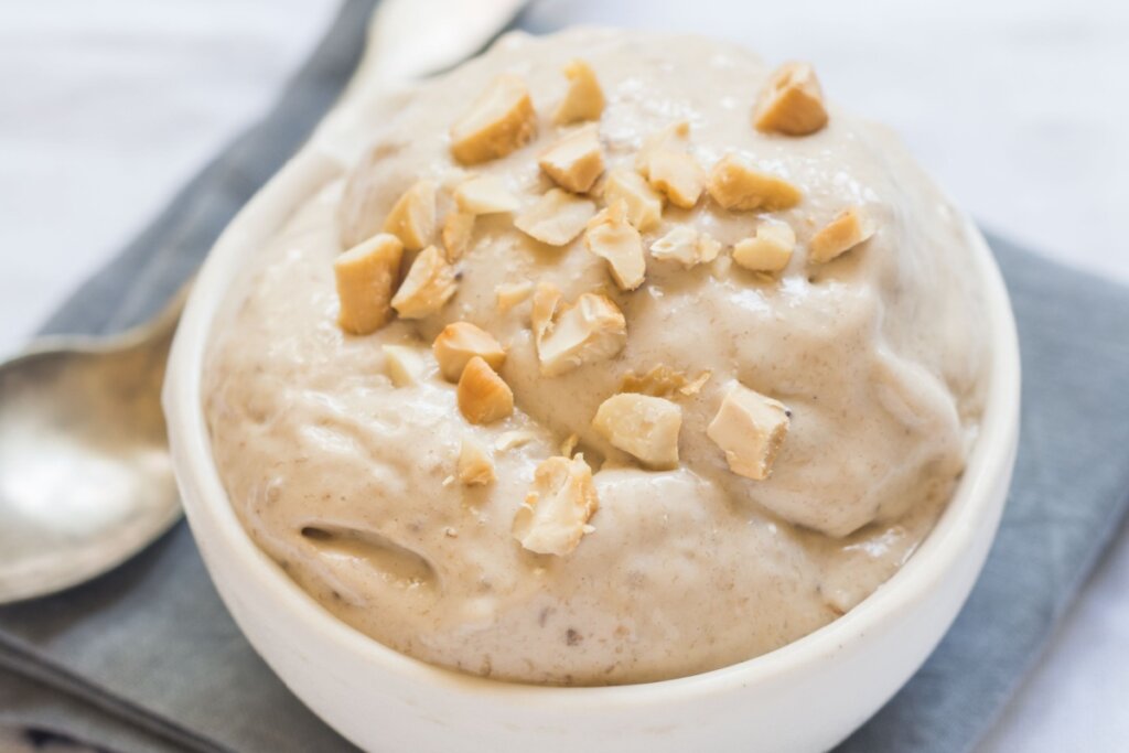pote branco com sorvete de banana e castanha-de-caju em cima sobre pano cinza com colher ao lado