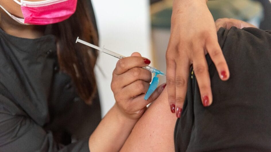 Foto mostra uma pessoa aplicando vacina em outra