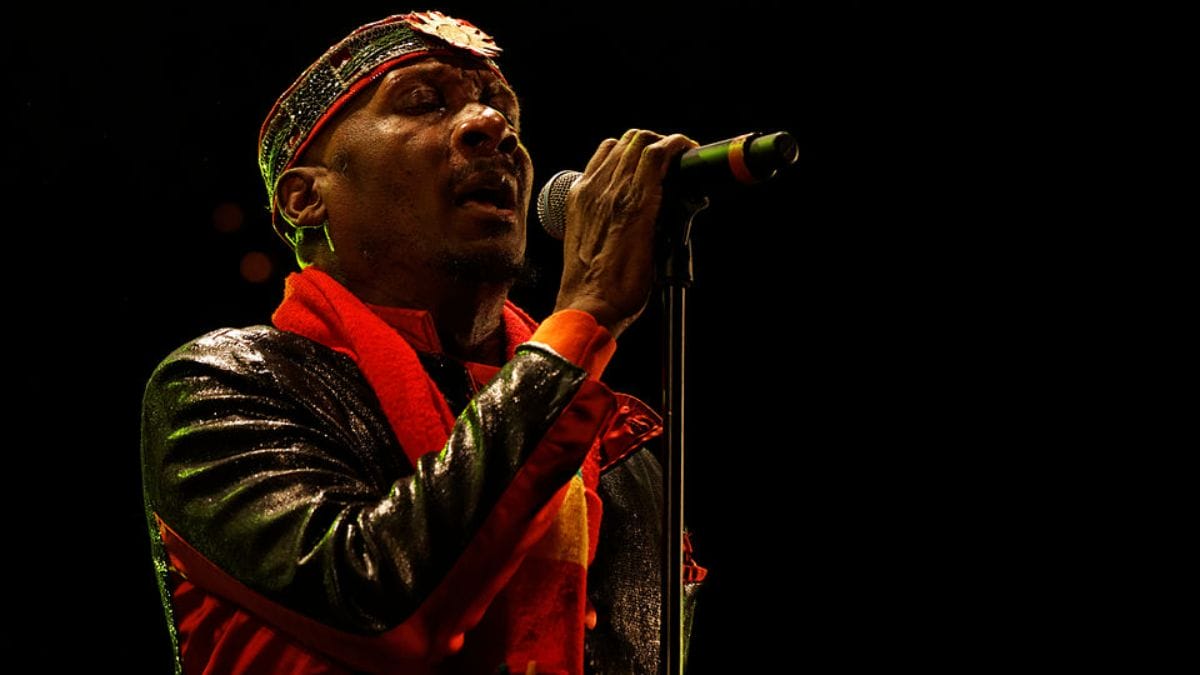 O cantor Jimmy Cliff sorrindo enquanto canta e toca violão em um show, simbolizando seu legado no reggae.