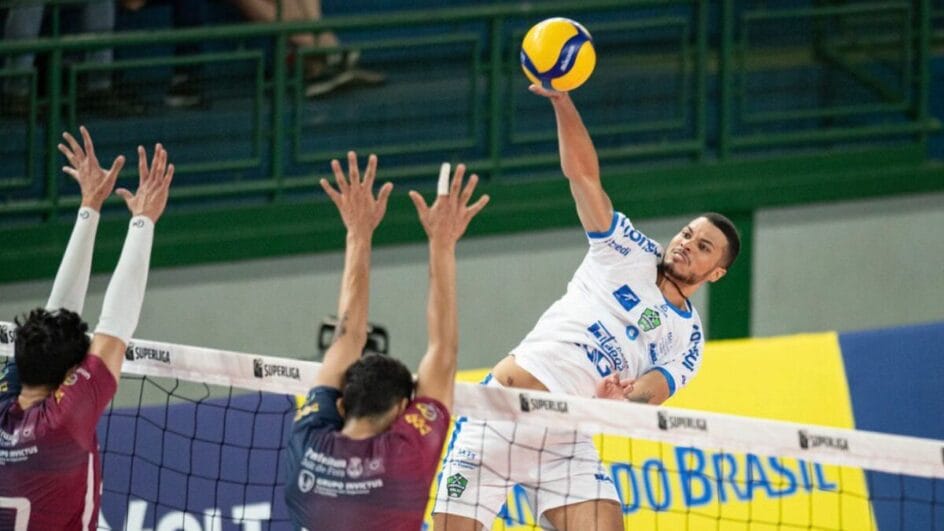 Atleta do Joinville vôlei atacando bola por cima da rede e com dois bloqeuadores na Superliga Masculina 202526