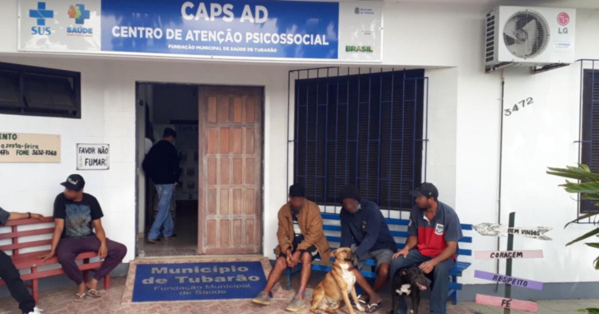 Fachada do CAPS em Tubarão, com pessoas aguardando sentadas. Os rostos das pessoas estão borrados