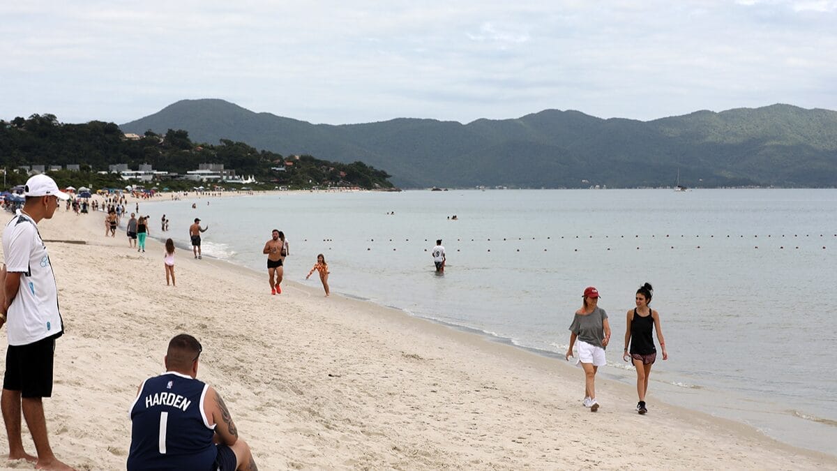 Imagem mostra a praia de Jurerê Internacional. O céu está nublado e há pessoas caminhando na orla da praia.