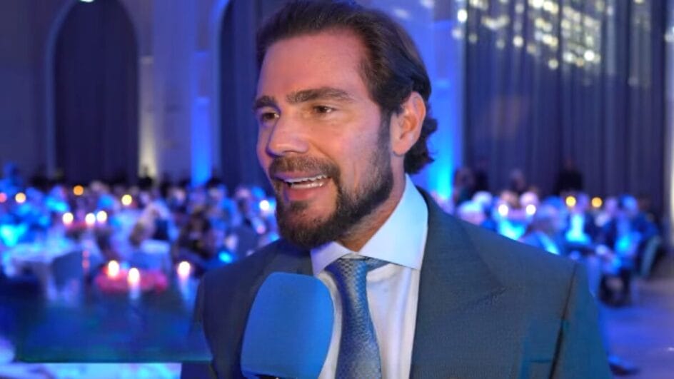 Daniel Vorcano dando uma entrevista em um evento. Ele usa terno da cor grafite com uma camisa branca. Daniel é branco, com cabelos marrons e barba.