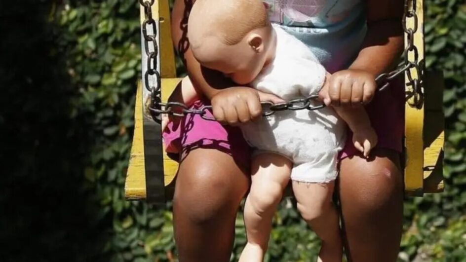 Foto mostra criança sentada em balanço segurando uma boneca