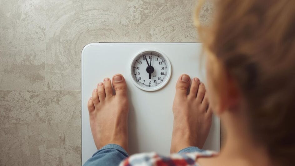 Mulher olhando para a balança com expressão de dúvida, ilustrando a variação de peso diária.