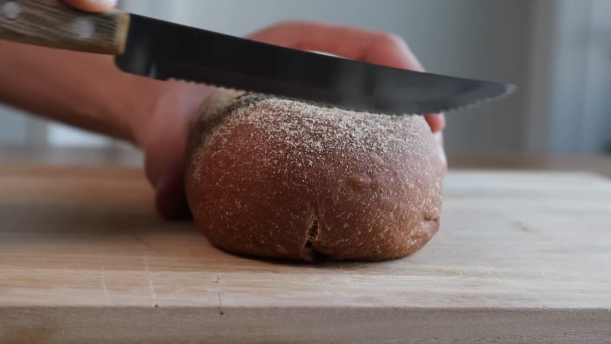 Pessoa com faca serrilhada cortando pão de coloração escura sobre tábua de madeira