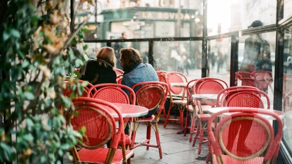 Pessoas sentadas em um restaurante. Cadeiras vazias de um lado e mesas ocupadas de outro. Dia de sol.