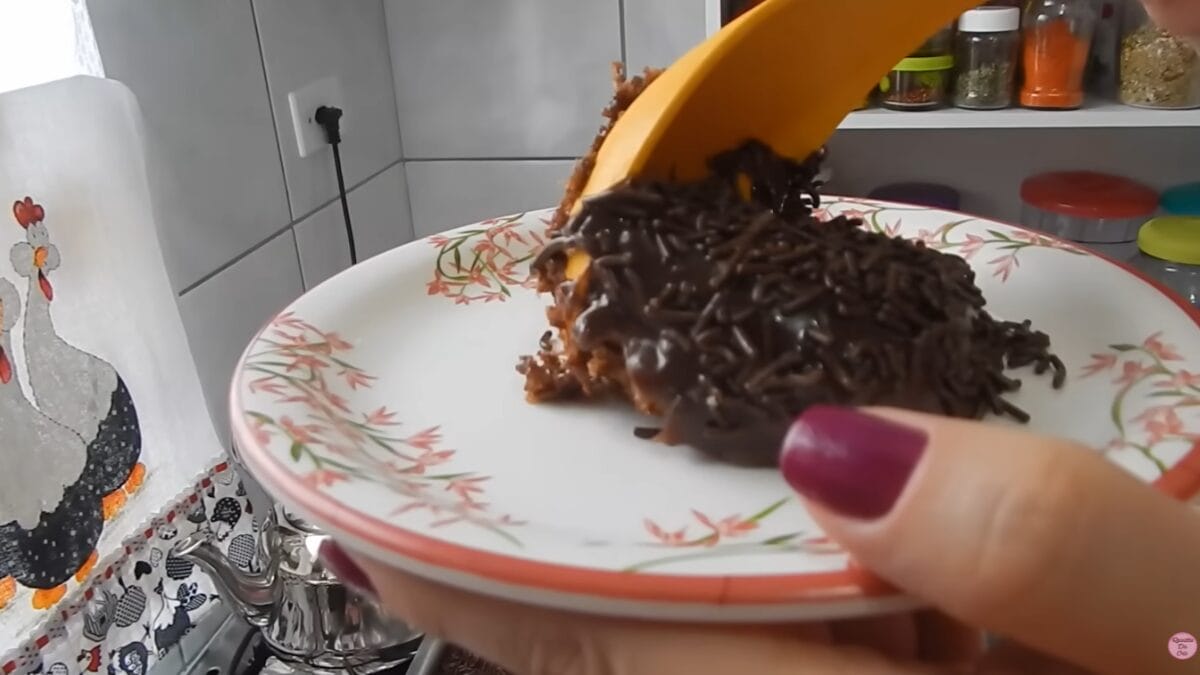 Fatia de bolo de chocolate com granulado sendo servida em prato floral em cozinha doméstica.