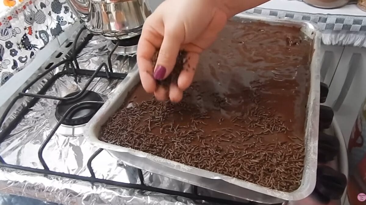 Mão espalhando granulado sobre cobertura de bolo de chocolate ainda quente na assadeira.