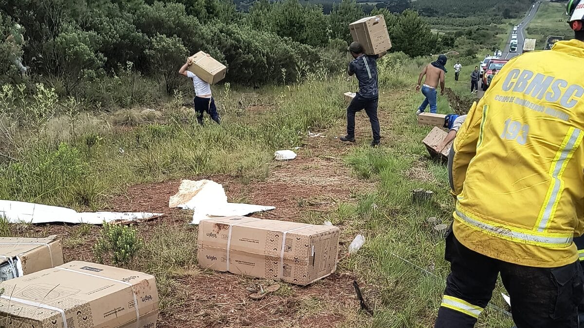 Pessoas pegam caixas da carga enquanto bombeiros atuam no caminhão tombado na quarta-feira (10) na BR-153, em Água Doce.