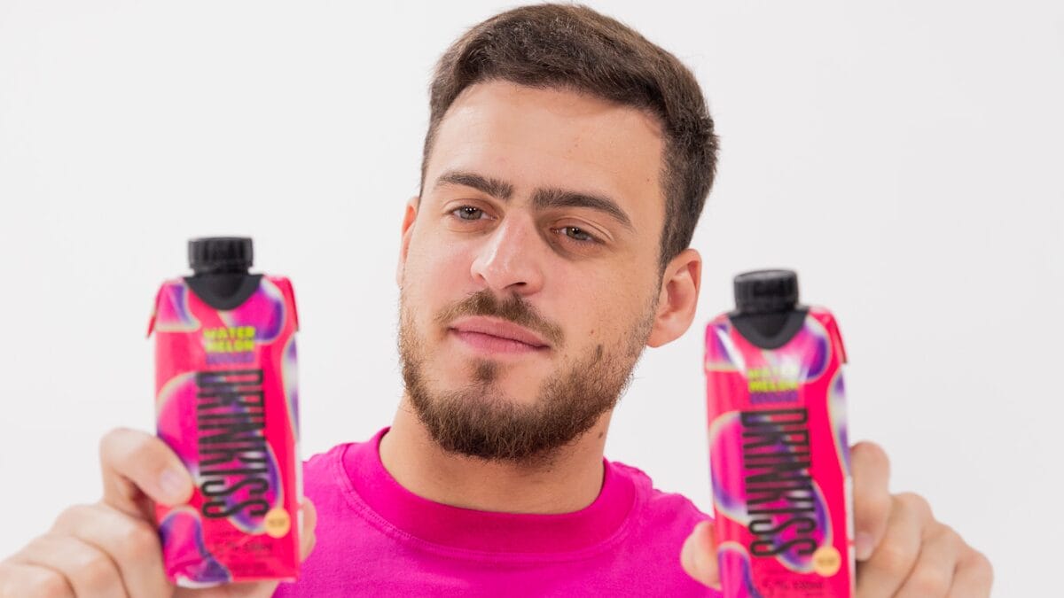 O influenciador Jon Vlogs, vestindo uma camiseta rosa vibrante, segura duas embalagens tetra pak na cor rosa da sua bebida "Drinkiss" sabor "Watermelon Sugar". Ele olha para a câmera com uma expressão séria contra um fundo branco neutro.
