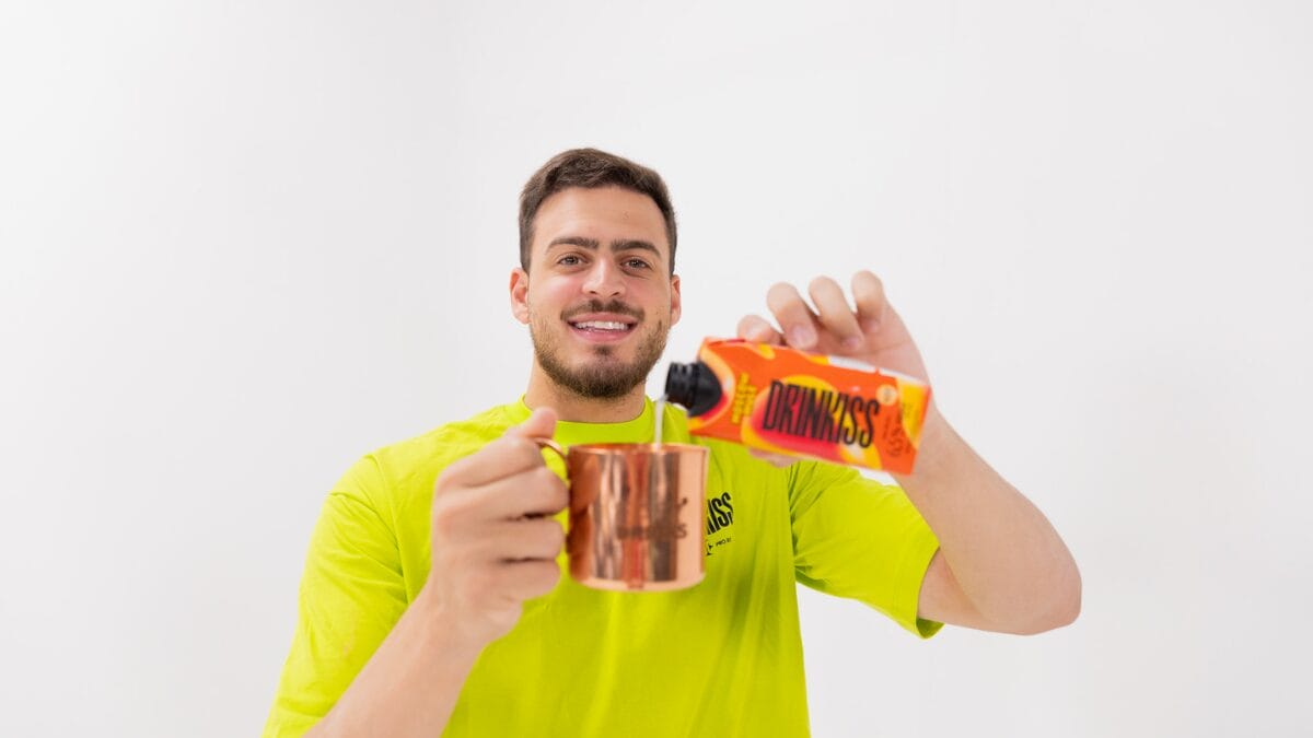 Jon Vlogs, usando uma camiseta verde-limão, sorri enquanto inclina uma caixa laranja de "Drinkiss Moscow Mule", servindo o líquido em uma caneca de cobre tradicional. O fundo é branco.