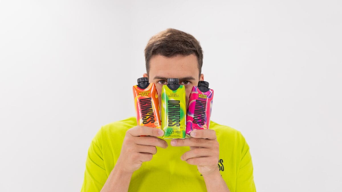 Jon Vlogs segura as três embalagens coloridas da Drinkiss juntas na frente do rosto, espiando por cima delas. As caixas são laranja (Moscow Mule), verde (Mate Tropical) e rosa (Watermelon Sugar).