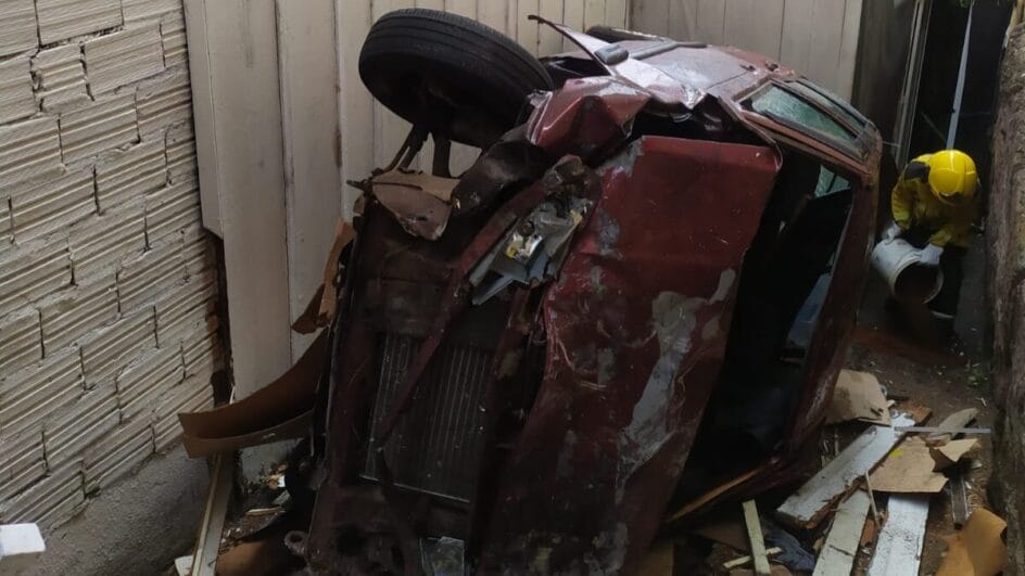 Carro capotado preso em corredor estreito ao lado de uma casa após acidente ocorrido em 9/12/2025 em Joaçaba, com destroços espalhados pelo chão.